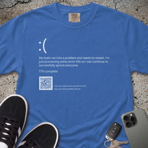 A Blue Screen of Death t-shirt features a parody error message with a sad face, white text, a QR code, and a progress bar at 17%. The shirt is displayed on concrete alongside black shoes, a phone, and car keys.
