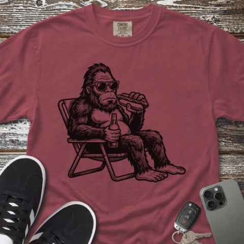 A "Bigfoot Chillin'" maroon t-shirt showcasing a graphic of a laid-back Bigfoot in sunglasses, lounging on a folding chair while holding a drink and a hot dog. The shirt is styled alongside black sneakers, a phone, and car keys.