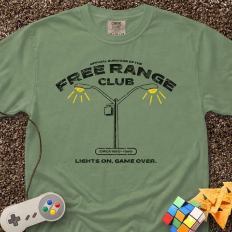 A green T-shirt reads "Free Range Club" with a streetlight graphic, "Circa 1970-1990," and "Lights On, Game Over." Surrounding items include a game controller, Rubik's Cube, tortilla chips, and a juice box.